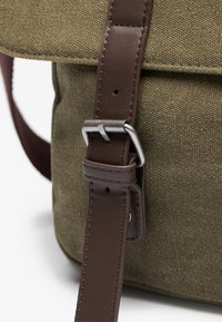 Olive green canvas bag with brown leather strap and metal buckle detail. Textured surface and clean lines characterize the design.