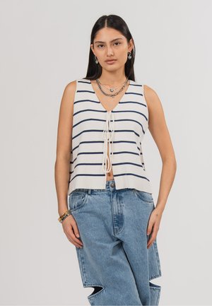 Frau mit langen dunklen Haaren, die ein ärmelloses weißes Oberteil mit marineblauen Streifen trägt, vorne gebunden, und weite blaue Jeans mit Ausschnitten an den Knien.