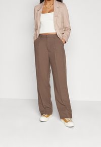 Blazer beige clair porté sur un crop top blanc, associé à un pantalon marron à jambes larges et des baskets blanches avec des accents jaunes. Tissu lisse.