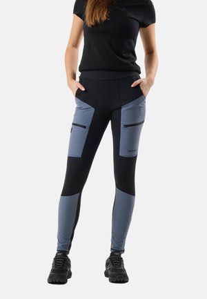 Vrouw draagt zwarte en grijs geblokte leggings met ritszakken op de dijen en zwarte sneakers, staat met handen in de zakken.