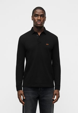 Άνδρας που φοράει μαύρο πικέ μπλουζάκι με μακριά μανίκια και μικρό πορτοκαλί λογότυπο "A|X" στο αριστερό στήθος, καθώς και σκούρο τζιν, στέκεται μπροστά σε απλό φόντο.