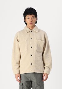 Cremefarbener Fleecejacke mit Knopfleiste, zwei Brusttaschen und Kragen. Verfügt über strukturierten Stoff und schwarze Knöpfe.
