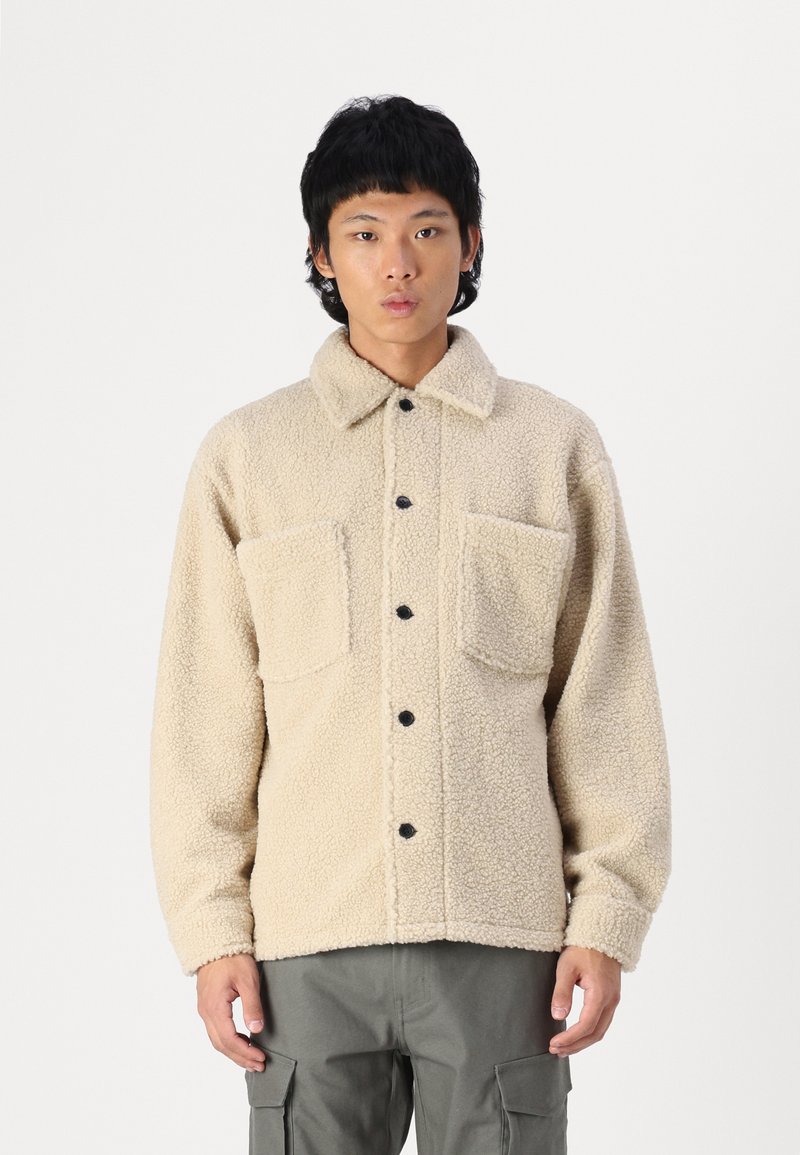 Cremefarbener Fleecejacke mit Knopfleiste, zwei Brusttaschen und Kragen. Verfügt über strukturierten Stoff und schwarze Knöpfe.