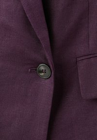 Gros plan sur un blazer violet foncé avec un bouton noir à quatre trous fermé et une petite poche sur le côté droit.
