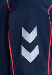 Ciemnoniebieska tkanina z dwoma białymi paskami w kształcie litery V i czerwonym obszyciem wzdłuż krawędzi, prawdopodobnie część rękawa odzieży sportowej.