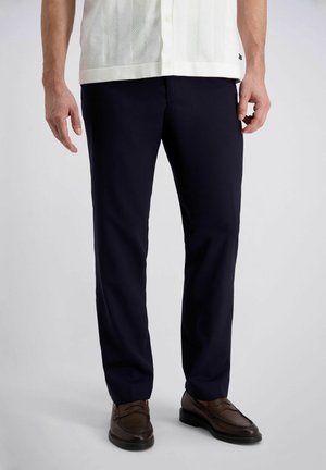 Mann trägt marineblaue Hose, weißes kurzärmeliges Hemd und braune Ledermokassins, steht vor weißem Hintergrund.