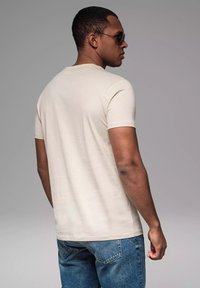 Beige kortærmet t-shirt med rund hals, båret med blå denim jeans. Model set forfra, iført mørke solbriller. Glat stoftekstur.