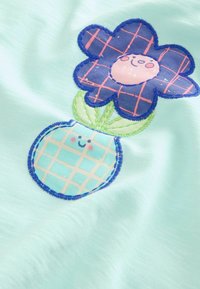 Fleur bleue souriante avec des pétales à carreaux roses et un visage rond au-dessus d'un pot rond bleu souriant avec un motif à carreaux roses sur un fond de tissu clair.