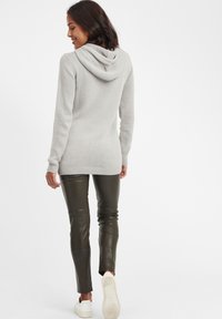 Hellgrauer gestrickter Hoodie in lockerer Passform, kombiniert mit dunkelgrünen Lederhosen und weißen Sneakern, von hinten betrachtet.