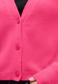 Cardigan tricoté rose vif avec boutons fermés sur le devant, porté sur un haut noir, main partiellement visible en bas.