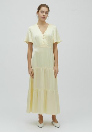 Kobieta stoi, ubrana w jasnofioletową sukienkę midi z falbanami, krótkimi rękawami, dekoltem w serek, guziczkami z przodu i białymi spiczastymi butami.