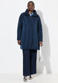 Cappotto impermeabile navy di lunghezza media con chiusura a zip, colletto alto e tasche laterali, abbinato a jeans scuri a gamba larga e stivali metallici alla caviglia.