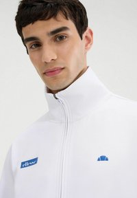 Chaqueta blanca con cremallera y cuello alto, que presenta un logo azul en el lado izquierdo del pecho y un pequeño detalle en azul. Textura de tela suave.
