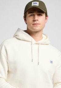 Sudadera blanca con bolsillo delantero y cordones, que presenta un pequeño parche de logo azul y blanco. Gorra verde oliva con visera plana.