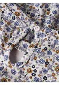 Mønstret stoff med små lilla, blå, oransje og svarte blomster på kremfarget bakgrunn; inkluderer rund hals med knappedetalj.