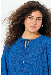 Blaue Bluse mit wirbelnden schwarzen Mustern, rundem Halsausschnitt und Knopfdetails, ausgestattet mit einer Schleife am Kragen und weichem Stofffaser.