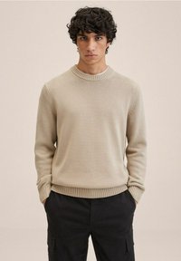 Beige gestrickter Pullover mit Rundhalsausschnitt, gerippten Bündchen und Saum, getragen über dunklen Cargo-Hosen. Glatte Textur, taillierte Silhouette.