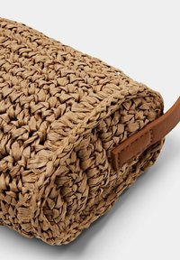Spleciona torba ze słomy w odcieniu tan z brązowym skórzanym paskiem. Zwróć uwagę na teksturowaną powierzchnię i okrągły kształt, podkreślający rzemieślniczy kunszt.