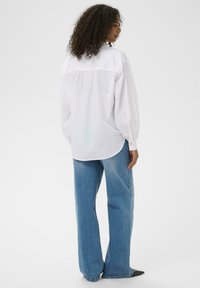 Une chemise blanche à manches longues avec une coupe décontractée, des plis à l'arrière et des poignets boutonnés, associée à un jean large bleu clair et des chaussures noires.