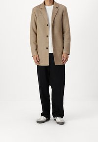 Beige uldblandingsfrakke med notched krave, enkeltknaplukning og struktureret overflade. Båret over en hvid ribbede sweater og sorte bukser.