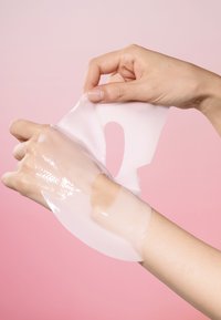 Masque en feuille clair, en gel, de couleur blanche, partiellement retiré d'une main. Texture lisse, conçu pour épouser la peau. Fond rose pâle.