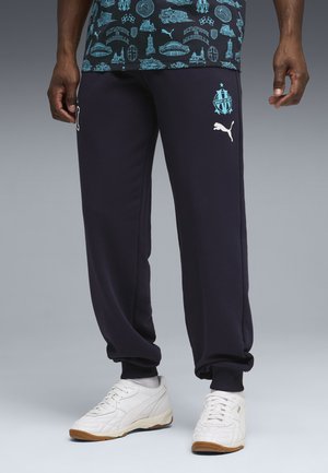 Marineblauwe sweatpants met geribbelde enkels, wit Puma-logo en embleem van Marseille. Draagt witte sneakers met een textuurontwerp.