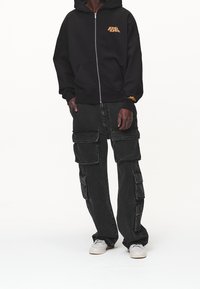 Felpa nera con zip e logo arancione, abbinata a pantaloni cargo neri larghi con più tasche e sneakers grigie.