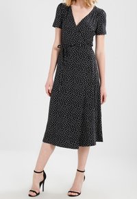 Czarna sukienka w stylu wrap z krótkimi rękawami, białe kropki i pasek w talii. Rozkloszowana midi o gładkiej fakturze.