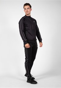 Zwarte sportieve trainingsoutfit bestaande uit een jack met rits en witte zijstrepen, en een broek met bijpassende strepen. Gemaakt van een gladde, elastische stof.