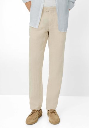 Uomo che indossa pantaloni di lino beige, camicia azzurro chiaro sbottonata e scarpe casual con lacci color cuoio su sfondo bianco.