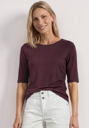 Femme aux cheveux blonds portant un haut violet foncé à manches courtes et un pantalon blanc, souriante sur un fond clair uni.