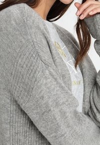 Grå stickad cardigan med ribbat mönster och långa, lösa ärmar, bärs över en ljusgrå skjorta med guldtryck.