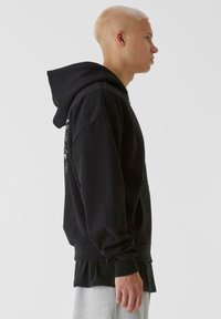 Svart huvtröja med framficka, ribbade mudd och fåll. Tyget verkar mjukt och har en lagerdesign. Slät textur, avslappnad stil.