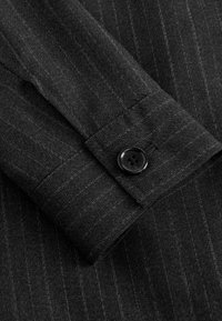 Manche de costume gris foncé à fines rayures avec un bouton noir à quatre trous sur le poignet, montrant une texture détaillée du tissu et des coutures.
