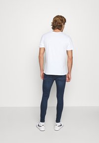 Camiseta de algodón blanca de mangas cortas, combinada con jeans ajustados de color azul oscuro. Etiqueta de marca visible en el bolsillo trasero. Usada con zapatillas blancas.