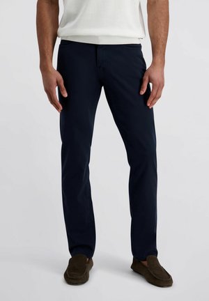 Man in marineblauwe slim-fit broek en bruine suède loafers, staand tegen een effen grijze achtergrond.