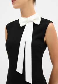Schwarzes Kleid mit einer weißen Satinschleife am Ausschnitt, ärmelloses Design und glatter Textur, das sich auf minimalistische Eleganz konzentriert.