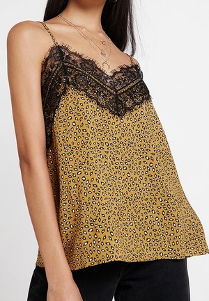 Top sans manches jaune moutarde pour femme avec bordure en dentelle noire et petit imprimé léopard noir et blanc, porté avec un pantalon noir.