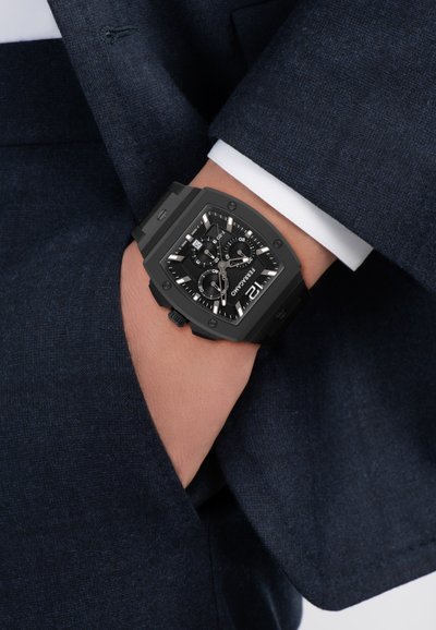 Na zápästí hodinky s čiernym štvorcovým chronografom, bielymi značkami a okienkom na dátum, v kombinácii s tmavomodrým oblekom a bielym manžetovým rukávom košele.