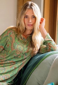 Blonde woman with loose hair wearing a green paisley shirt leans on a green cushion on a couch near a window.