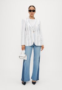 Striped white blazer with button closure, paired with light blue flared jeans. Model holds a small white handbag and wears black sunglasses.