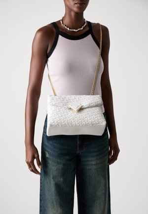 Woman wearing a white sleeveless top and denim jeans with a white woven handbag featuring a gold chain strap across her chest.