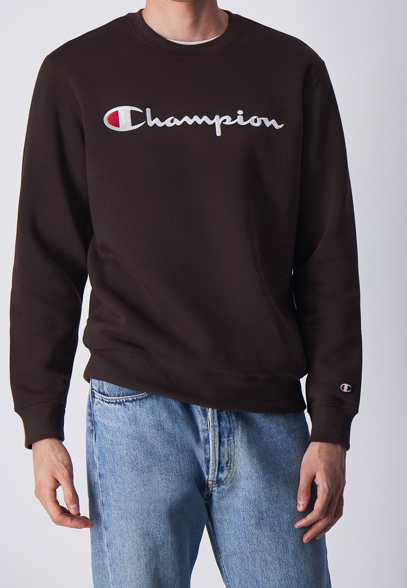 Felpa marrone in misto cotone, con logo "Champion" ricamato in bianco e piccolo accento logo sulla manica sinistra. Indossati jeans blu.