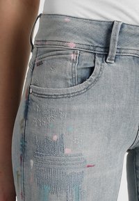 Ljust blå denimjeans med hög midja, som har en blandning av broderade och målade mönster i rosa, blått och konstnärliga design.