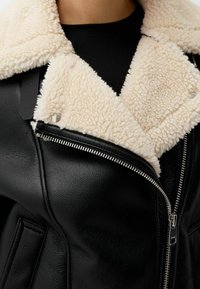 Giubbotto di pelle nera con colletto in morbido shearling crema. Presenta una zip argentata e tasche piatte, evidenziando una superficie liscia e strutturata.