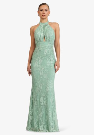 Frau trägt ein mintgrünes, bodenlanges Spitzenkleid mit Neckholder und einem vorderen Schlüsselloch-Detail, steht vor einem weißen Hintergrund.