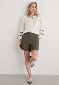 Vit långärmad skjorta med blå och röda mönstrade detaljer, olivgröna shorts och beige vävda sandaler på en neutral bakgrund.