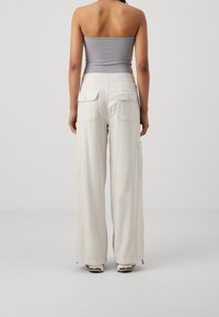 Široké cargo kalhoty ve světle béžové barvě s dvěma zadními kapsami. Hladká, lehká struktura látky. Modelka má na sobě šedý top bez ramínek.