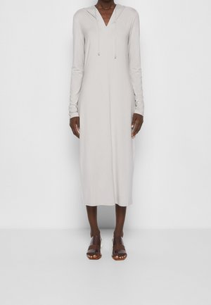 Mujer con vestido midi de manga larga en gris claro, con escote en V y capucha, acompañada de sandalias marrón oscuro de punta abierta, posando frente a un fondo blanco.