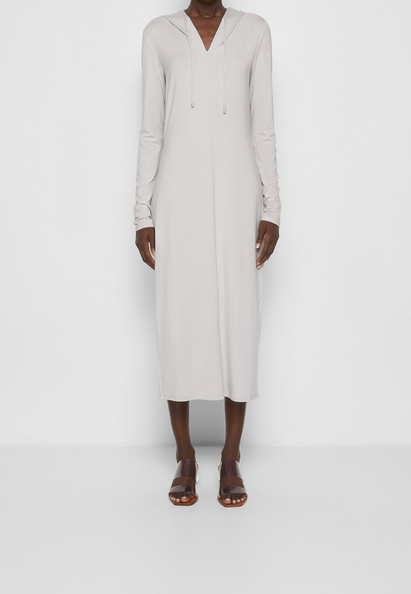 Femme portant une robe midi gris clair à manches longues avec un col en V et une capuche, associée à des sandales à bout ouvert couleur marron foncé, debout sur un fond blanc.
